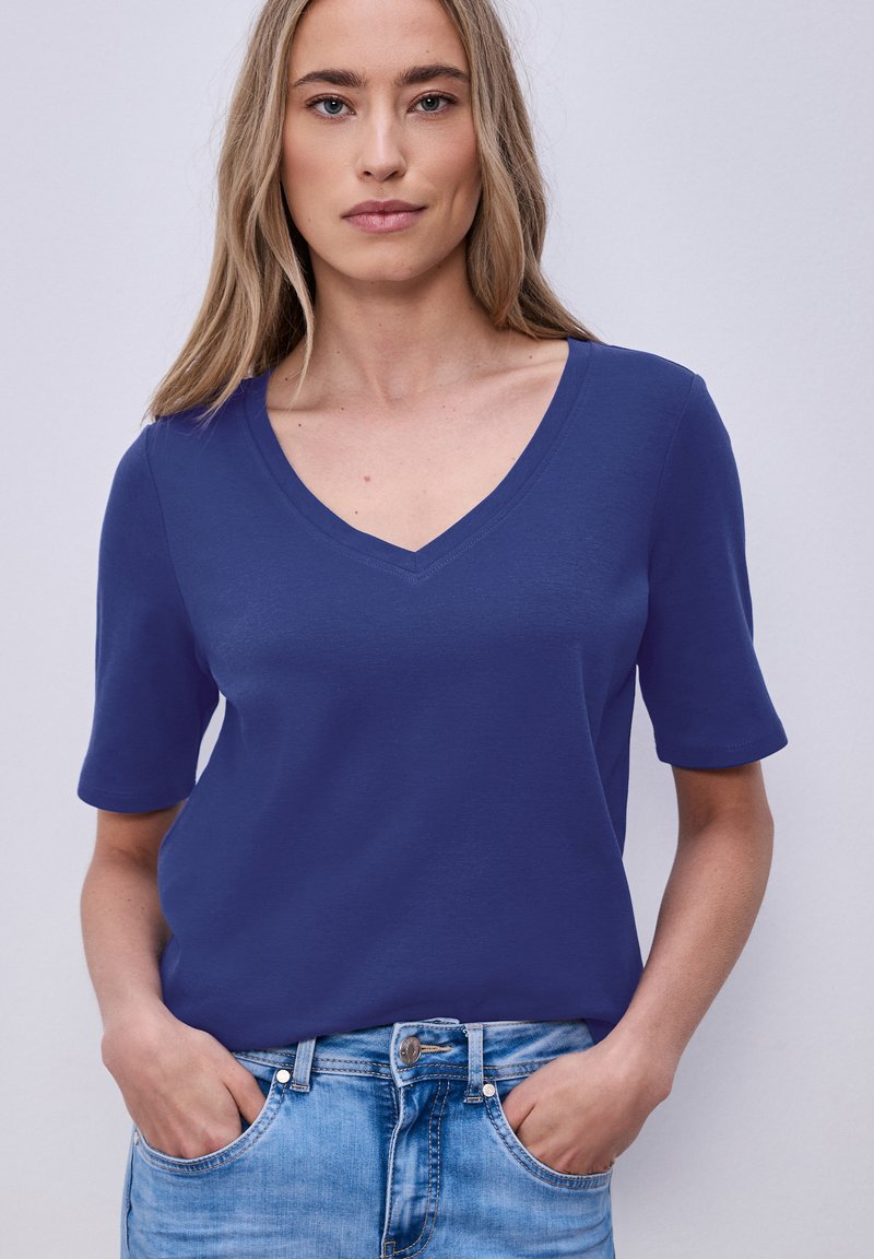 Vrouw draagt een blauw T-shirt met V-hals en korte mouwen, ingestopt in lichtblauwe spijkerbroek, met handen in de zakken tegen een effen achtergrond.
