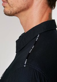 Camisa negra con un tejido texturizado, cuello puntiagudo y un acento en la costura lateral con el logotipo de "CAMP DAVID" en blanco.