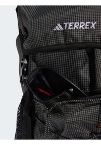 Black backpack with grid pattern, featuring a front pocket holding sunglasses. Material is textured, with adjustable straps and metal hardware.
