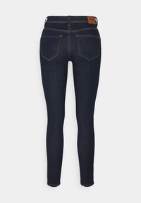 Vaqueros skinny azul oscuro con bolsillos traseros, lazos para cinturón y una etiqueta de marca de cuero marrón en la cinturilla, visto desde atrás.