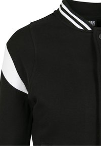 Veste universitaire noire avec une bande blanche sur la manche, col côtelé, boutons-pression et texture lisse. Présente un design épuré et sportif.