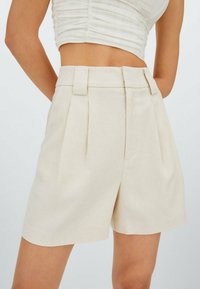 Shorts plissés beiges en tissu léger, dotés d'une taille ajustée avec des accents matériels subtils et une texture lisse.