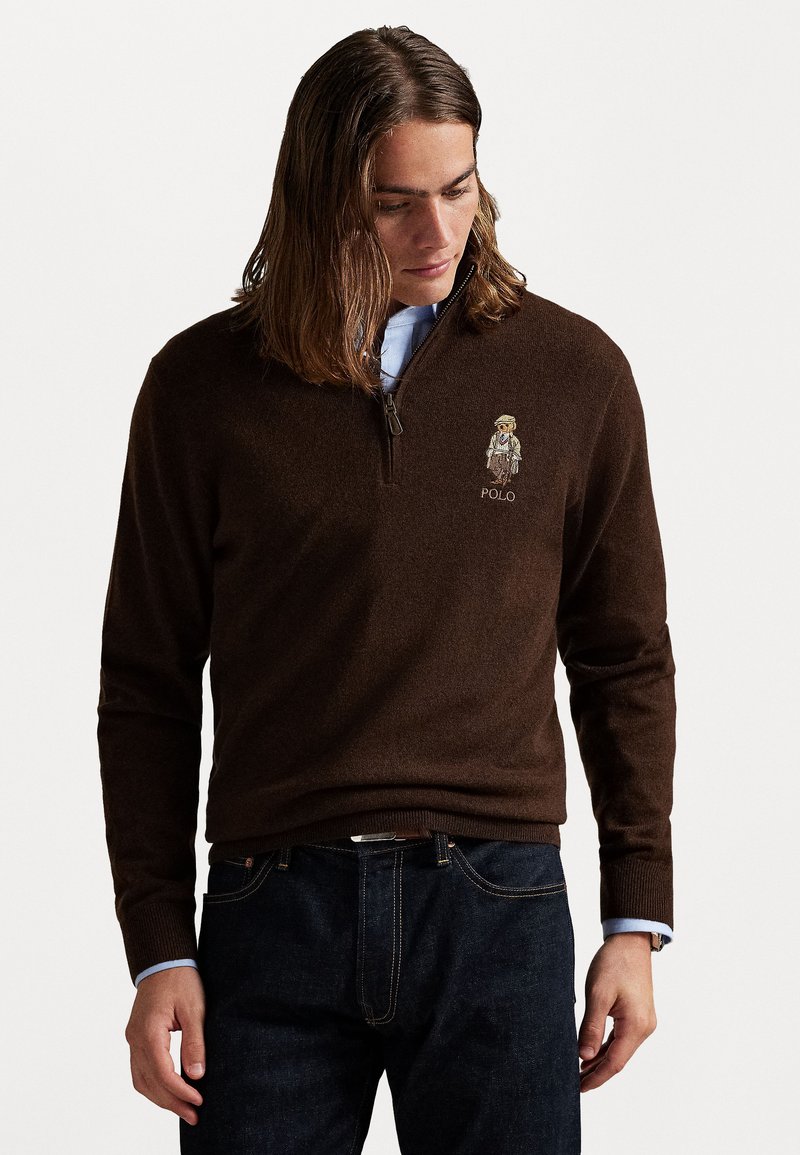 Rjava zadrgo pulover iz mehkega blaga, z vezenim motivom in besedilom "POLO" na prsih, v kombinaciji s temnimi kavbojkami.