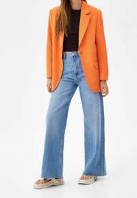 Kvinna i en ljusorange blazer, svart topp, högmidjade vida ljusblå jeans och beige plattformssandaler mot vit bakgrund.