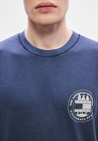 Tamsiai mėlynas medvilninis marškinėlis su apvalia iškirpte, trumpomis rankovėmis ir kairėje krūtinės pusėje esančiu apvaliu logotipu su lauko tema susijusiais grafika.