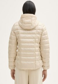 Veste sans manches beige avec un design matelassé horizontal, capuche et poignets élastiques. Texture lisse et matériau léger.