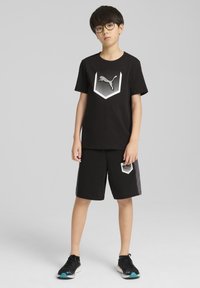 T-shirt di cotone nero con logo esagonale bianco e nero, abbinato a pantaloni corti neri con logo corrispondente. Sneakers nere con dettagli teal.