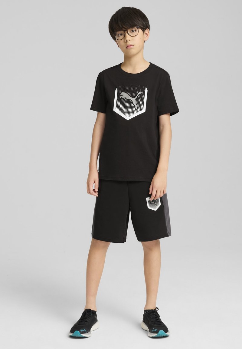 T-shirt di cotone nero con logo esagonale bianco e nero, abbinato a pantaloni corti neri con logo corrispondente. Sneakers nere con dettagli teal.