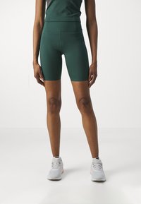 Høyt liv, mørkegrønne treningsshorts i et glatt, elastisk stoff som slutter midt på låret. Kombinert med lysegrå joggesko.