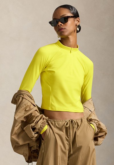 Λαμπερό κίτρινο μακρυμάνικο τοπ με φερμουάρ στο ένα τέταρτο, συνδυασμένο με φαρδιά καφέ παντελόνια. Ένα ανοιχτό καφέ φαρδύ μπουφάν κρέμεται από το ένα χέρι.