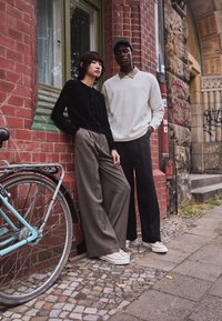 Two models stand against a brick wall. One wears a black cardigan and gray wide-leg trousers; the other wears a white sweater and black pants.