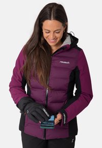 Lila und schwarze isolierte Jacke, mit Reißverschluss, graue Innenseite, getragen mit schwarzen Handschuhen und hält ein Smartphone. Reusch-Branding sichtbar.