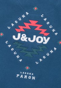 Blauwe stof met geborduurde tekst en geometrische motieven. Bevat witte en rode accenten, met zigzagontwerpen en decoratieve patronen.