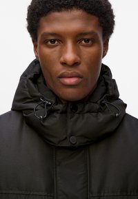 Chaqueta puff negra con cuello alto, ajustes con cordones y cierres de botones. Exterior liso con una textura sutil.