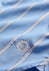 Světle modrá pruhovaná košile s bílými a tmavě modrými pruhy, zdobená vyšívaným kruhovým logem. Měkký bavlněný materiál s hladkou texturou.