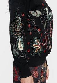 Maglione nero con intricati ricami floreali nelle tonalità di verde, rosso e oro, con paillettes e cuciture dettagliate sulle maniche.