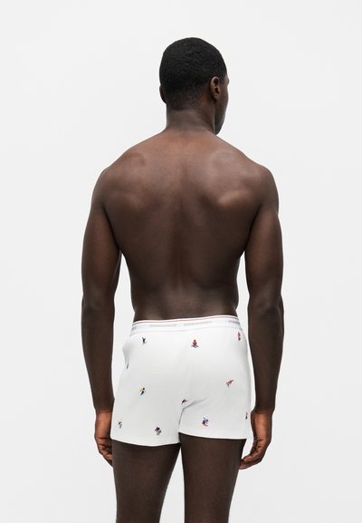 Homme musclé à la peau foncée, debout, dos tourné, portant un boxer blanc avec de petites impressions colorées de joueurs sportifs sur un fond uni.