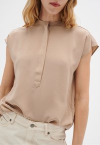 Beige blus med hög halsringning, korta ärmar och en subtil knäppning. Mjuk, slät tyg utan synliga mönster eller detaljer.