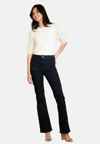 Donna in piedi che indossa un top con maniche in pizzo bianco, jeans neri a vita alta e sandali con tacco a blocco beige, sorridente con una mano sulla coscia.