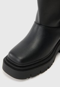 Botim de tornozelo em couro preto com biqueira quadrada, acabamento texturizado, sola robusta preta e detalhes discretos com nervuras ao longo da borda.