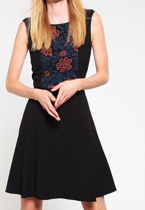 Frau mit langen blonden Haaren, die ein ärmelloses schwarzes Kleid mit roter und blauer Blumenstickerei auf der Brust trägt, Hände vorne verschränkt.