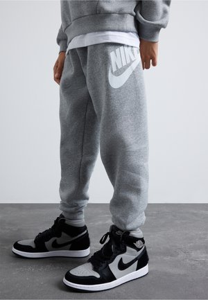 Personne portant un pantalon de survêtement gris Nike et des baskets montantes Nike noires et blanches debout sur un sol gris.
