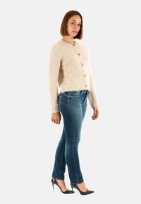 Maglione beige e peloso con collo rotondo e cinque bottoni, abbinato a jeans blu attillati e décolleté nere. Trama liscia.