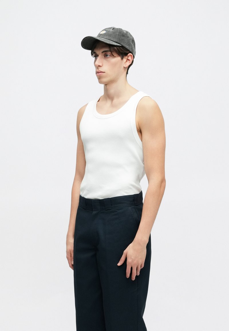 Weißes geripptes Tanktop kombiniert mit dunklen Hosen und einer grauen Kappe. Die Kappe hat ein gesticktes Design und einen gebogenen Schirm.