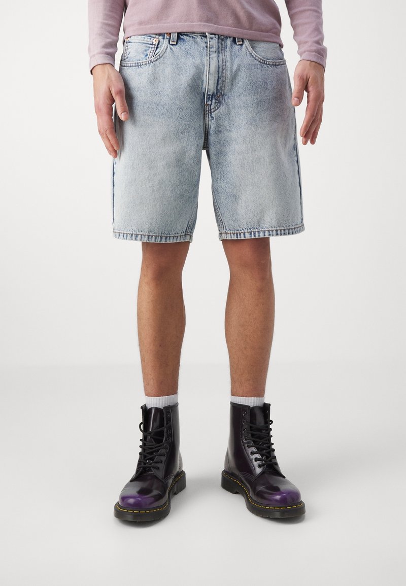 Shorts en denim bleu clair à coupe décontractée, avec poches latérales et ourlets effilochés, associés à des bottes noires à lacets ornées d'accents violets et de coutures jaunes.