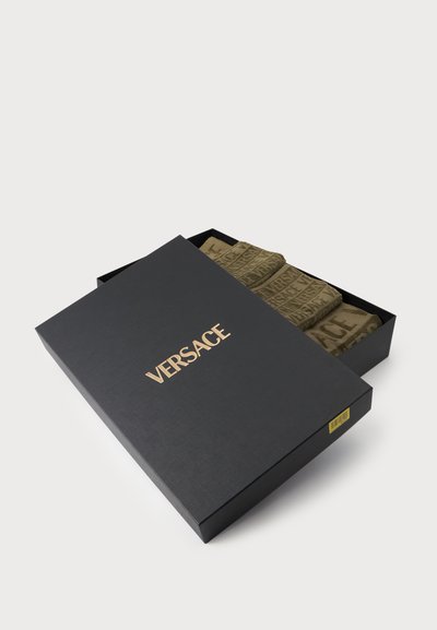 Μαύρο κουτί με το χρυσό λογότυπο VERSACE, μερικώς ανοιχτό ώστε να αποκαλύπτει διπλωμένα υφασμάτινα αντικείμενα σε λαδί χρώμα με σχέδιο το κείμενο VERSACE στο εσωτερικό.