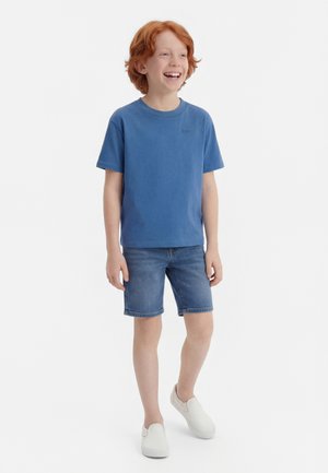 Smilende rødhåret dreng, stående iført en blå t-shirt, denimshorts og hvide slip-on sko mod en ensfarvet hvid baggrund.