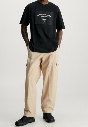 Sort bomulds T-shirt med et frontgrafisk logo i guld, parret med beige cargo bukser og sorte sko. Casual og afslappet pasform.