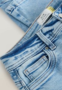 Kitsad keskmise kõrghuse denim teksad helesinisena, millel on pleekinud plaastrid, kontrastne õmblus ja väike eesmine tasku. Silt, kus on tootmisdetailid, on nähtav.