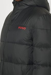 Schwarze Steppjacke mit horizontalem Steppmuster, hoch geschlossenem Kragen, Reißverschluss vorne und rotem "HUGO"-Logo auf der linken Brustseite.