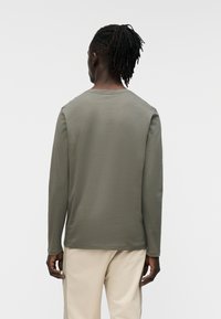 Μακρυμάνικο πουκάμισο σε ελαιόχρωμο πράσινο χρώμα με λαιμόκοψη τύπου crew, απαλή υφή και ευθεία ραφή στο τελείωμα, συνδυασμένο με ανοιχτό μπεζ παντελόνι.