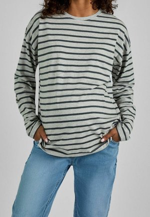 Person iført en grå langærmet skjorte med sorte vandrette striber og lyseblå jeans, hænderne i lommerne mod en ensfarvet baggrund.