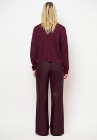 Blonde vrouw in een bordeaux gebreide trui en bijpassende brede leren broek, staande tegen een vlakke witte achtergrond.