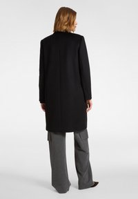 Cappotto di lana nera con silhouette dritta, maniche lunghe e taglio a metà. Indossato sopra pantaloni cargo grigi a gamba larga.