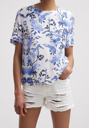 Top blanco con estampado floral azul, mangas cortas, escote redondo. Combinado con shorts de denim blancos desgastados con bordes deshilachados.