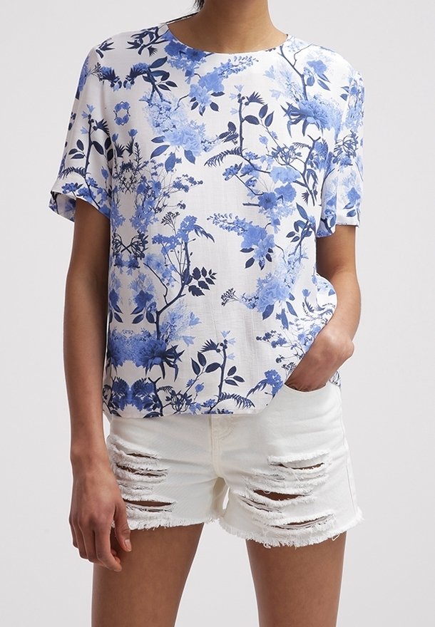 Top blanco con estampado floral azul, mangas cortas, escote redondo. Combinado con shorts de denim blancos desgastados con bordes deshilachados.