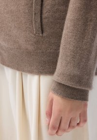 Brun ulltröja med ribbad nederkant och ärmslut, med en framficka, parat med vita strukturerade byxor. Närbild på ärm och hand.