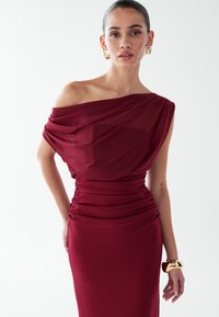 Vīnogu krāsas off-shoulder kleita ar drapētām auduma detaļām, rūpīgi darināta jostas daļā un pieguļoša silueta. Akcentējošs zelta aproce.