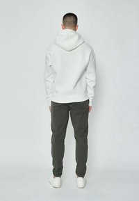 Homme de dos portant un hoodie blanc, un pantalon sombre et des baskets blanches devant un fond clair.