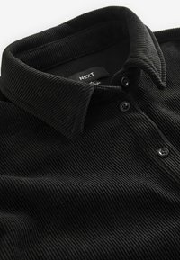 Schwarzes Cord-Polohemd mit strukturiertem Kragen, drei Knöpfen und einem Etikett mit der Aufschrift "NEXT." Die Textur ist gerippt.