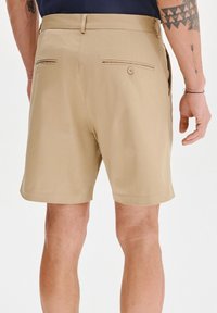 Beżowe szorty wykonane z gładkiego materiału, z dwiema tylne kieszeniami zapinanymi na guziki oraz prostym wykończeniem. Brak widocznych wzorów lub akcentów.