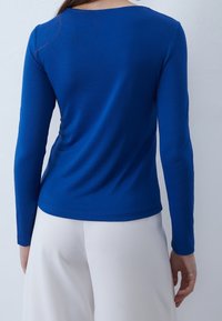 Maglia blu a maniche lunghe realizzata in tessuto liscio, con scollatura tonda e forma aderente, abbinata a pantaloni chiari.