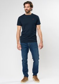 Tumed sinine puuvillane t-särk ümmarguse kaelusega ja väikese punase logoga, kombineeritud siniste teksade ja pruunide nahast tossudega.