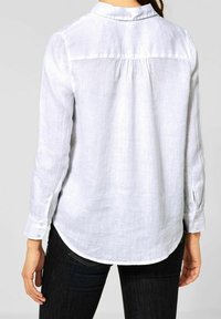 White linen shirt with a collar, long sleeves, and button cuffs. Features a pleated back design and curved hem. Textured fabric visible.