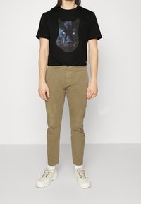 Hombre que lleva una camiseta negra con una gran imagen de la cara de un puma negro, pantalones caqui, calcetines blancos y zapatillas beige, de pie contra una pared lisa.