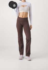 Atletický outfit zahrnující světle béžové tričko s dlouhým rukávem a výstřihem, hnědé obtažené kalhoty a světle růžové sportovní boty. Závaží v levé ruce.